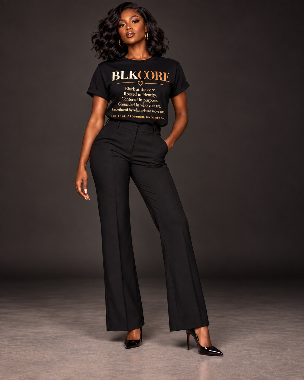 A woman modeling a black short-sleeve graphic T-shirt with gold text reading 'BLKCORE' and a definition-style print, paired with black wide-leg pants and black heels, against a dark studio background.