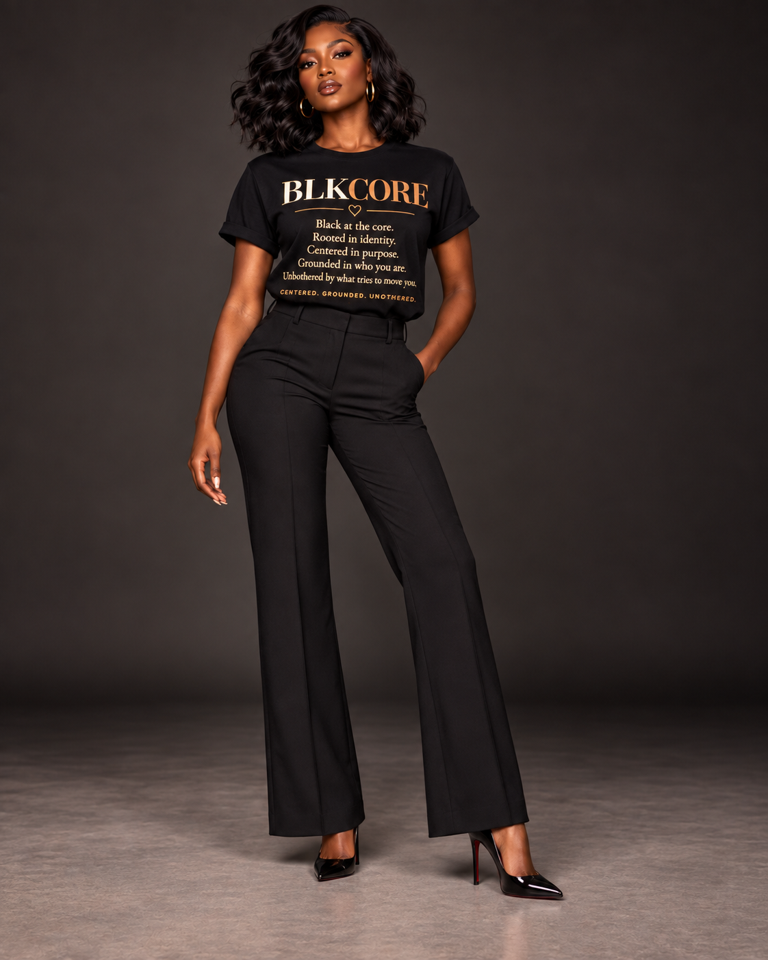A woman modeling a black short-sleeve graphic T-shirt with gold text reading 'BLKCORE' and a definition-style print, paired with black wide-leg pants and black heels, against a dark studio background.