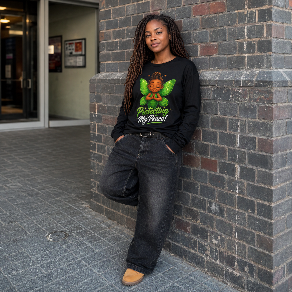 A woman with long dreadlocks stands against a gray brick wall wearing a black long-sleeve graphic t-shirt with a colorful angel design and the text 'Protecting My Peace!'.