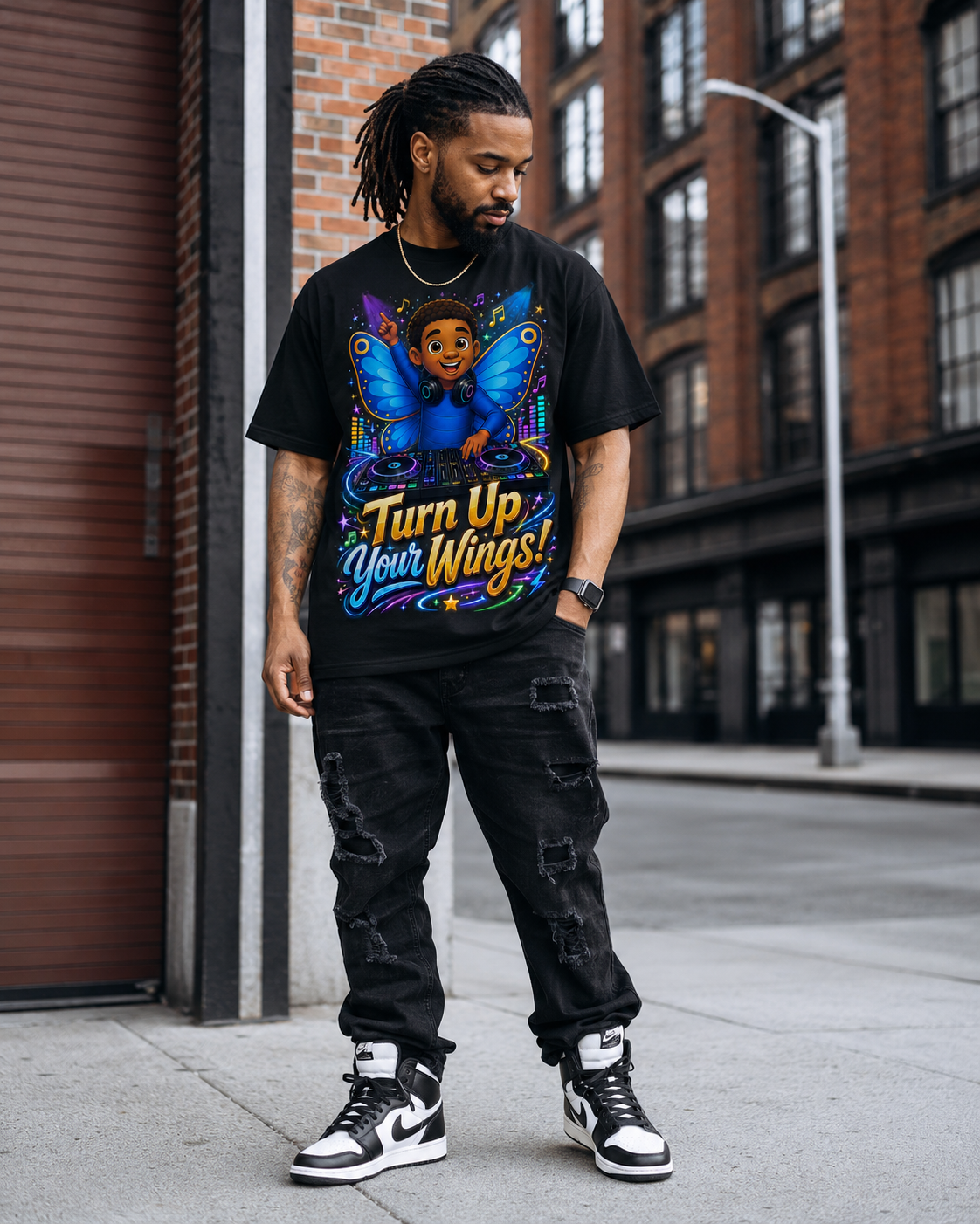 A man wearing a black graphic t-shirt with a colorful DJ winged angel design and the text 'Turn Up Your Wings!' paired with distressed black jeans and black-and-white sneakers, standing on a city sidewalk.