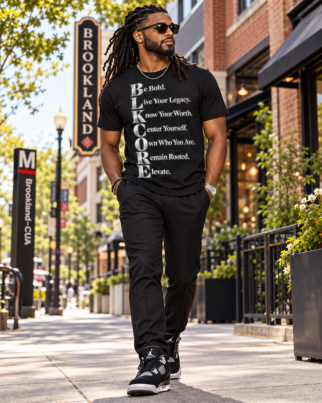 A man wearing a black graphic t-shirt with the text 'BE BOLD, See Your Legacy, Know Your Worth, Center Yourself, Trust Who You Are, remain Rooted, and Believe.' walks down a sunny city street.