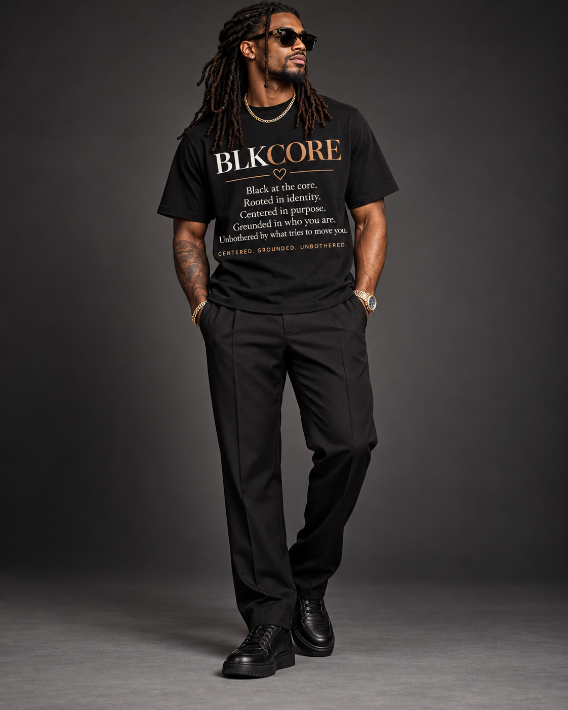 A man modeling a black graphic t-shirt with 'BLK CORE' printed in gold and white text, paired with black trousers and black shoes, set against a dark gray studio background.