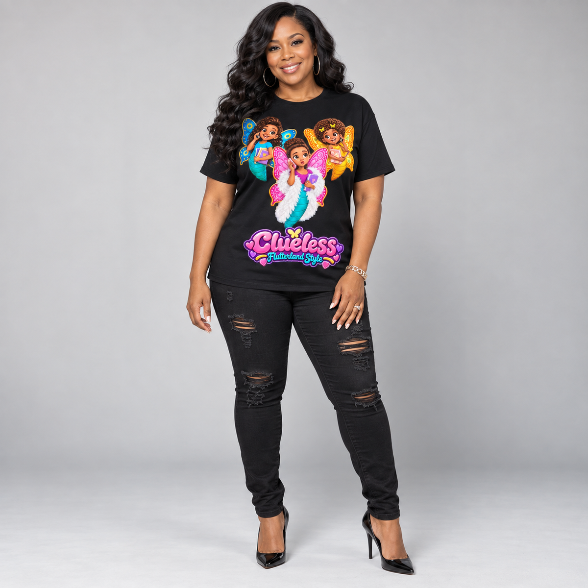 A woman modeling a black graphic t-shirt with colorful cartoon characters and the text 'Clueless Remember Me', paired with black distressed skinny jeans and black high heels, against a light gray studio background.