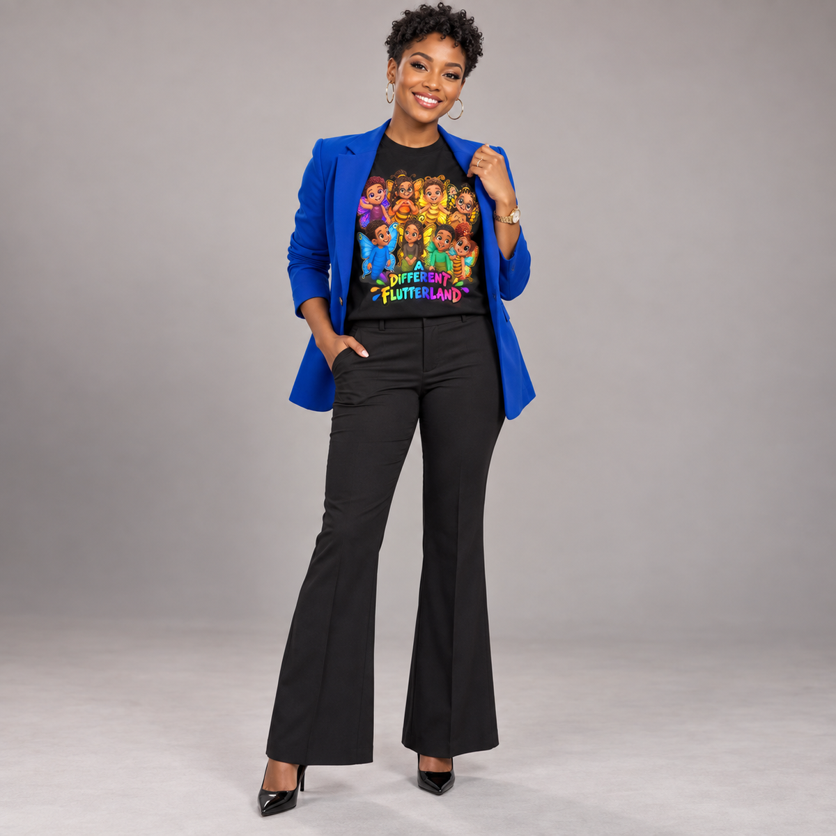 A woman stands against a light gray studio background wearing a bright blue blazer, black flared pants, and a black graphic T-shirt with a colorful cartoon group print and the words 'WITCHES OF FUTTLELAND'.