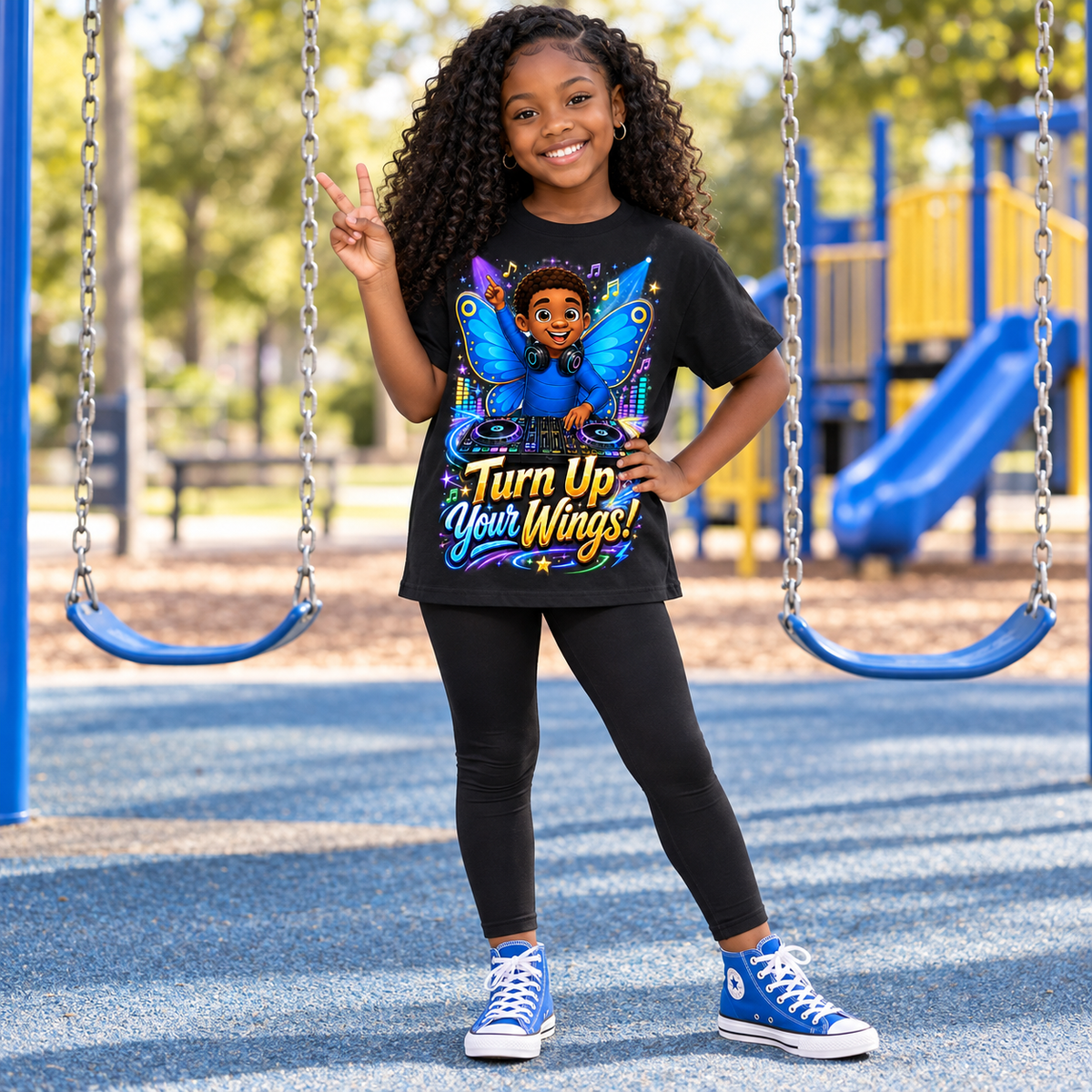 A young girl with curly hair stands on a playground wearing a black graphic t-shirt with a colorful butterfly design and the text 'Turn Up Your Wings!'. She is also wearing black leggings and blue sneakers.