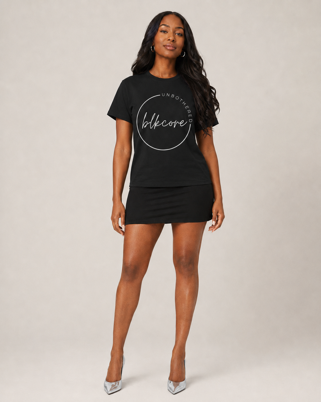 A woman modeling a black short-sleeve t-shirt dress with a white circular graphic on the front that reads 'UNBOTHER blckcore', paired with a matching black mini skirt and silver high heels, against a neutral studio background.