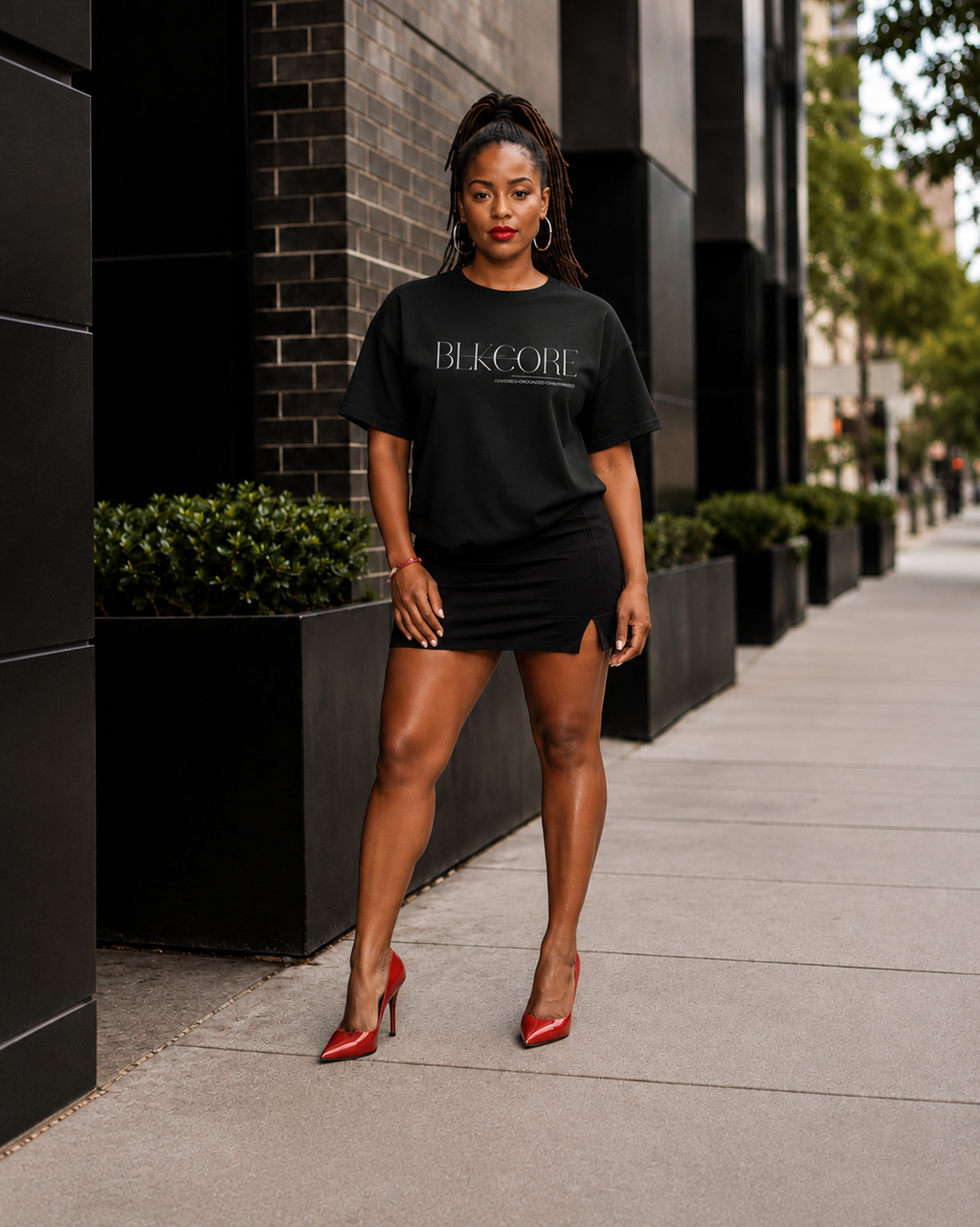 A woman stands on a city sidewalk wearing a black oversized T-shirt dress with 'BLKCORE' printed in white across the chest, paired with red pointed-toe heels. The background features a modern black brick building and planters.