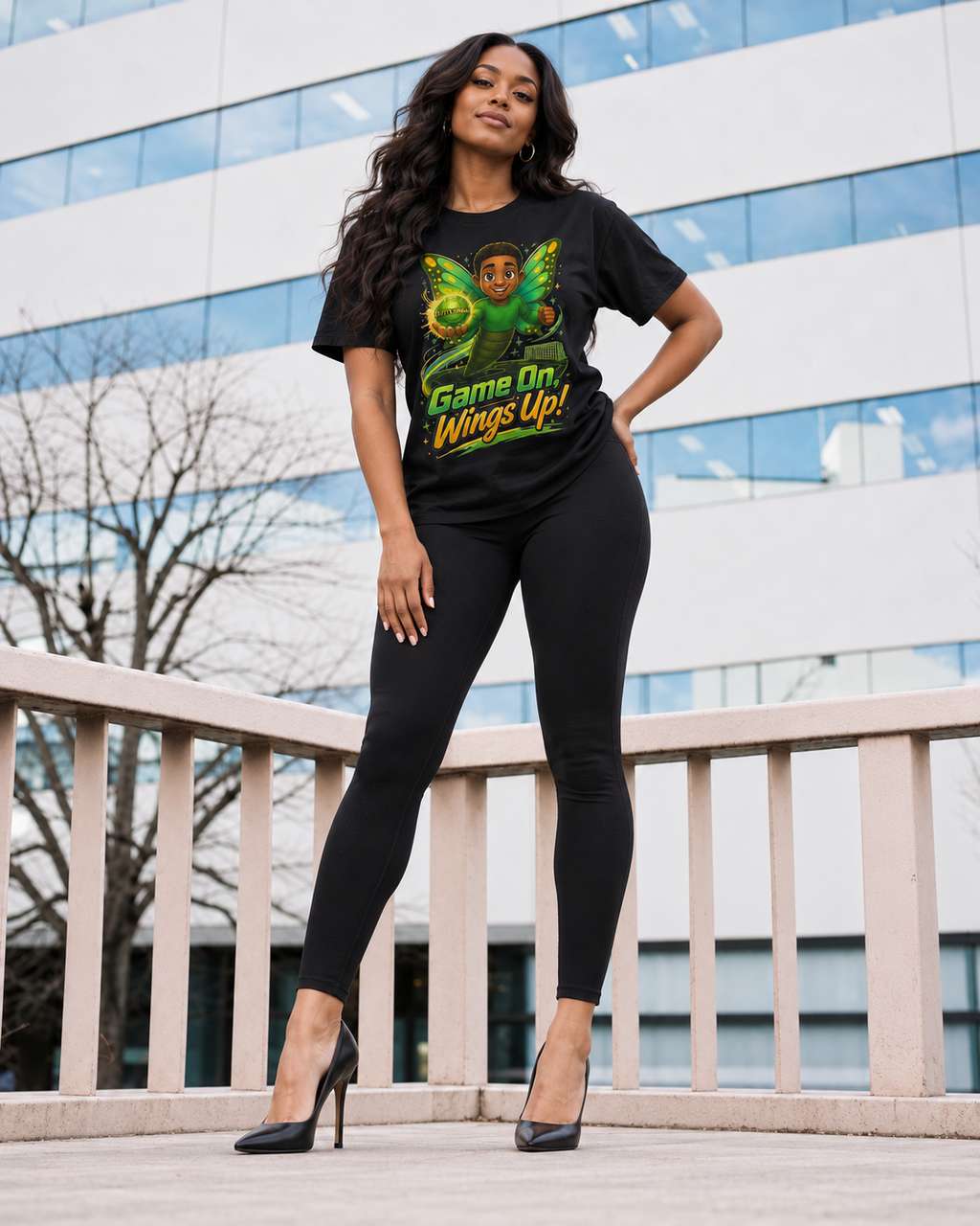 A woman stands outdoors in front of a modern glass building, wearing a black graphic T-shirt with a green butterfly and the words 'Game On Wings Up!' along with black leggings and black high heels.