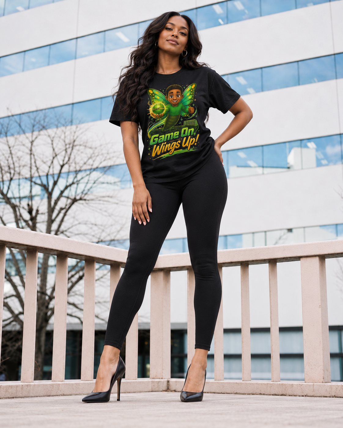 A woman stands outdoors in front of a modern glass building, wearing a black graphic T-shirt with a green butterfly and the words 'Game On Wings Up!' along with black leggings and black high heels.